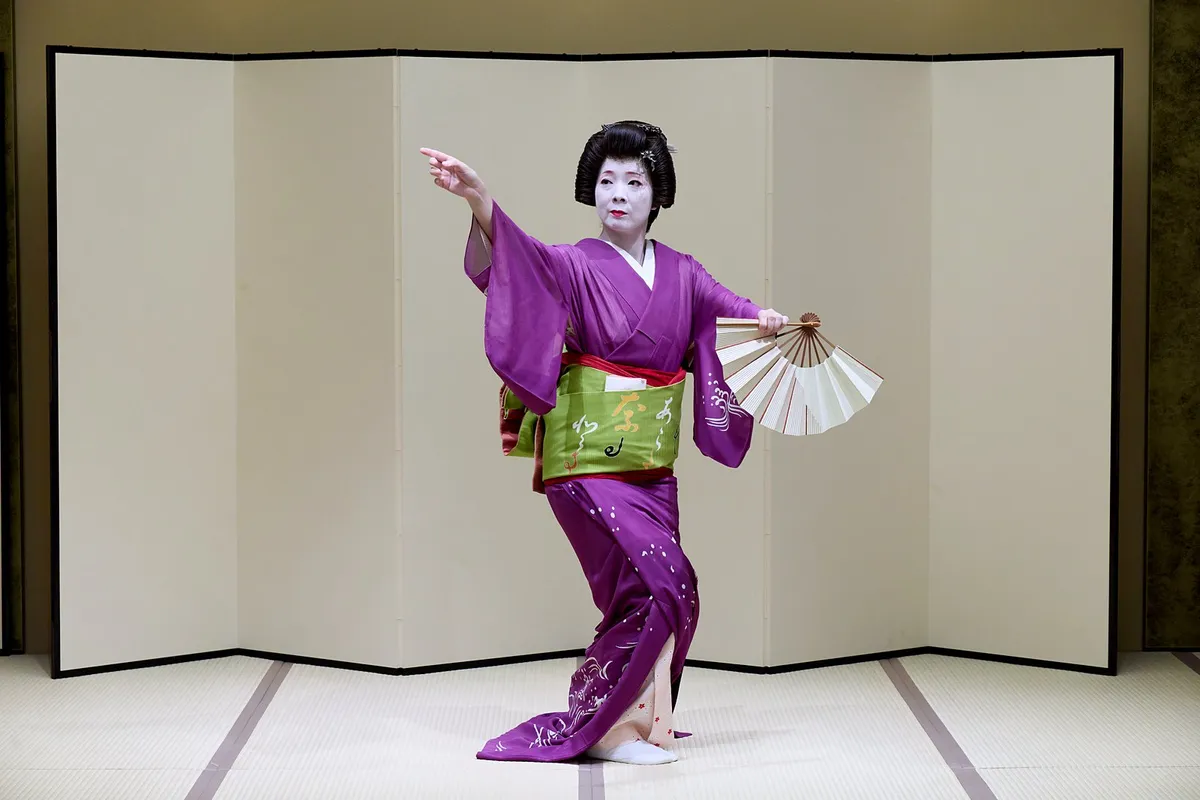 屏風の前で、片手を差し伸べ、もう片方の手で扇を開いて踊る浅草の芸者。