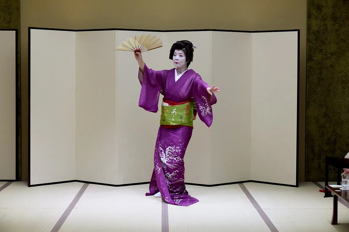 紫の着物の裾を畳に引いて、扇を掲げて踊る立方の芸者。