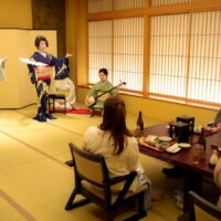 浅草 都鳥のお座敷。屏風の前で芸者衆2名が扇を手に舞い、三味線に合わせてお客様が膳を前に見ている