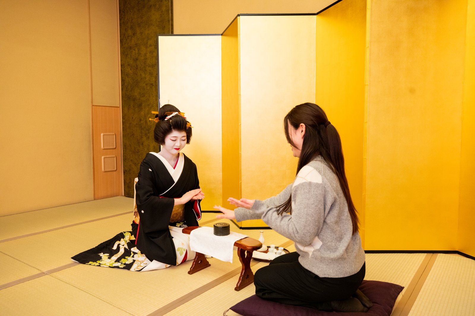 金屏風の前で、芸者とお客様が赤い木の台をはさんでお座敷遊び「こんぴらふねふね」をしている